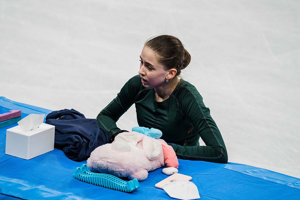 A figure skater resting rink-side beside the ice, leaning on a table with a plush toy and personal items after a performance.
