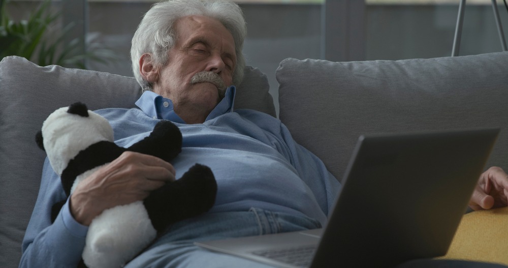 Elderly man resting on a sofa while holding a panda plush toy, with a laptop open beside him, conveying comfort and companionship.
