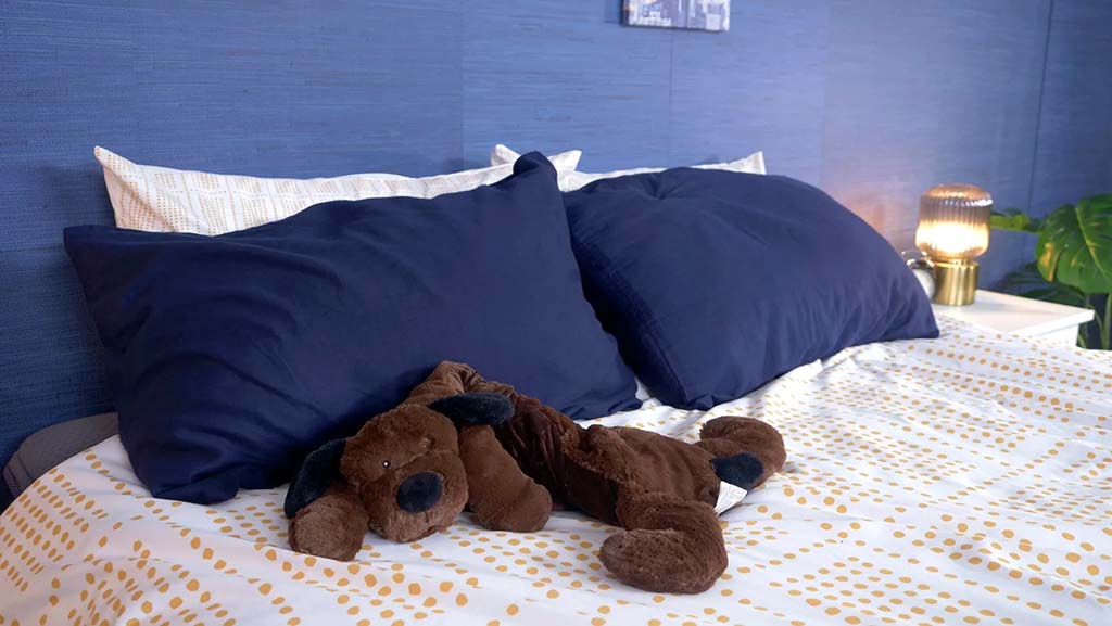 Brown plush puppy toy lying flat on a neatly made bed with white and gold polka-dot bedding, dark blue pillows, and a soft blue wall, creating a calm and cozy bedroom atmosphere.