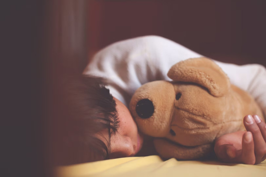 Young child sleeping on a bed while hugging a soft teddy bear close to their face in a warm, low-light bedroom.