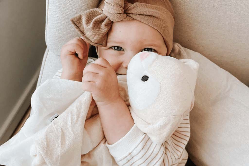 Smiling baby cuddling a soft white plush toy while resting on a cushioned chair, conveying comfort, security, and gentle bonding.