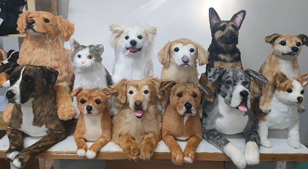 Group display of realistic handmade pet replica plush toys, featuring multiple dog breeds and a cat, arranged together on a workshop table to showcase likeness accuracy and fur detailing.
