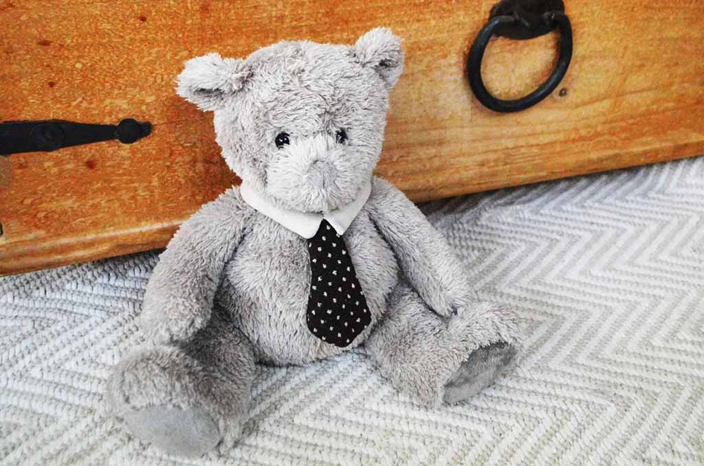 Gray plush teddy bear wearing a white collar and black polka-dot tie, seated on a textured fabric surface in front of a wooden chest, with a soft, slightly worn appearance that suggests a classic or vintage style.