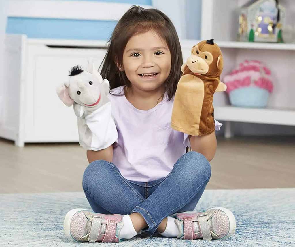 Smiling child sitting cross-legged on the floor, holding two soft hand puppets—a light gray animal puppet in one hand and a brown monkey puppet in the other—inside a bright, cozy playroom.