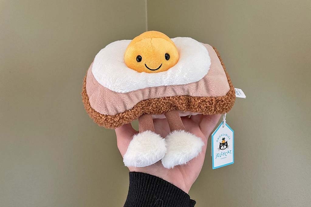 Hand holding a Jellycat fried egg on toast plush toy with smiling yolk, fluffy white egg, textured brown toast base, dangling legs, and visible Jellycat brand tag.