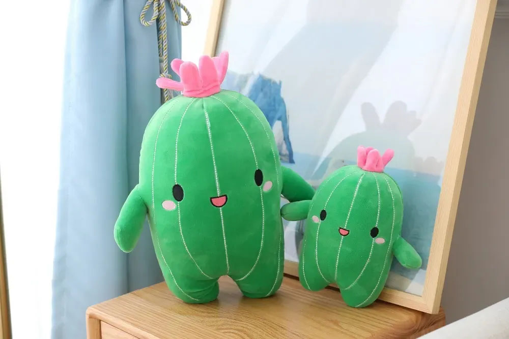 Cute cactus plush toys in two sizes standing on a wooden table, featuring smiling faces, stitched white lines, and pink flower tops in a cozy home setting.