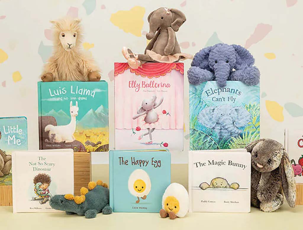 A Jellycat plush toy display featuring an elephant, llama, bunny, and egg characters placed alongside colorful children’s picture books, arranged neatly against a soft pastel background.