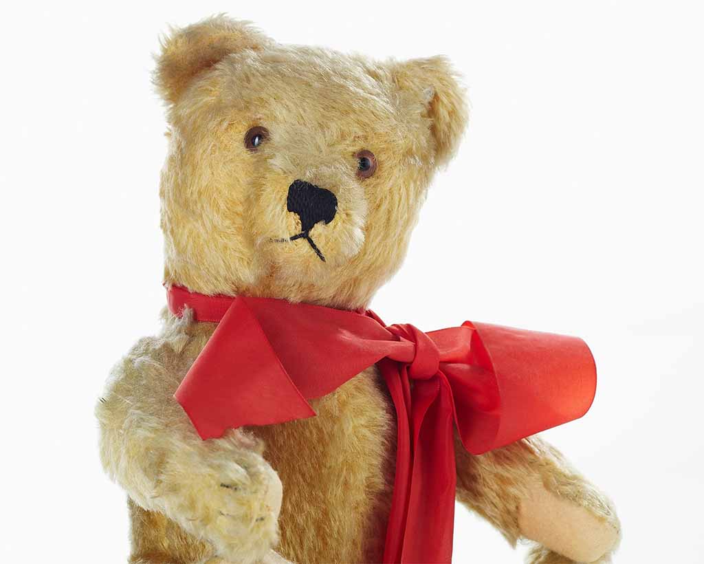 Vintage-style teddy bear with light brown fur wearing a red ribbon bow, photographed against a white background.