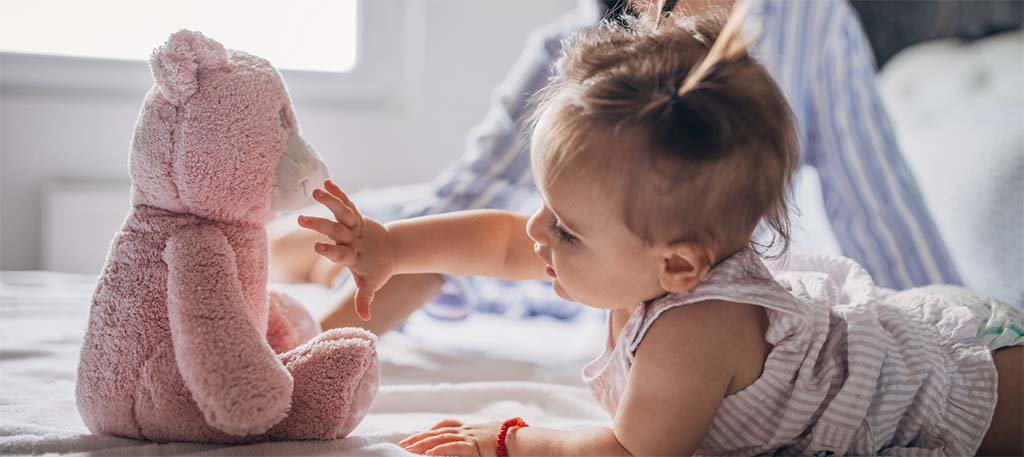Baby lying on a bed reaching out to touch a soft pink teddy bear, showing gentle interaction and early sensory exploration.