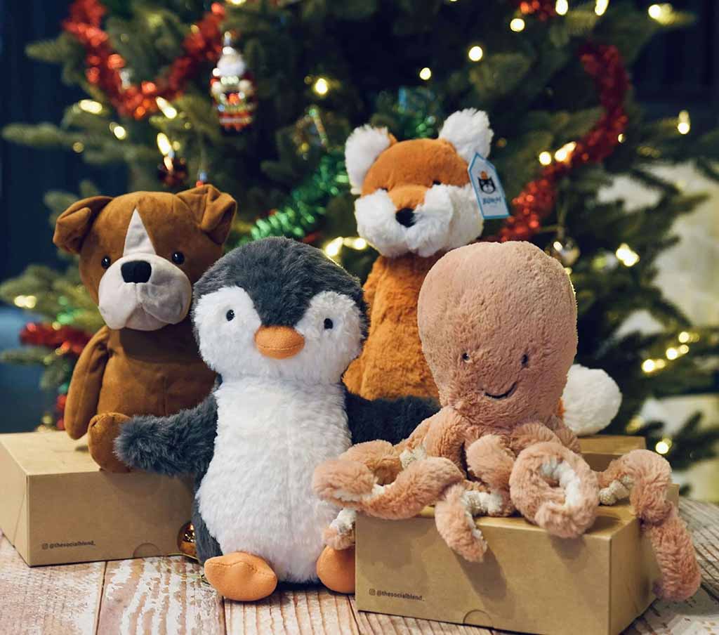 Four Jellycat plush toys—including a dog, penguin, fox, and octopus—sitting on gift boxes in front of a decorated Christmas tree with warm lights and festive garlands.