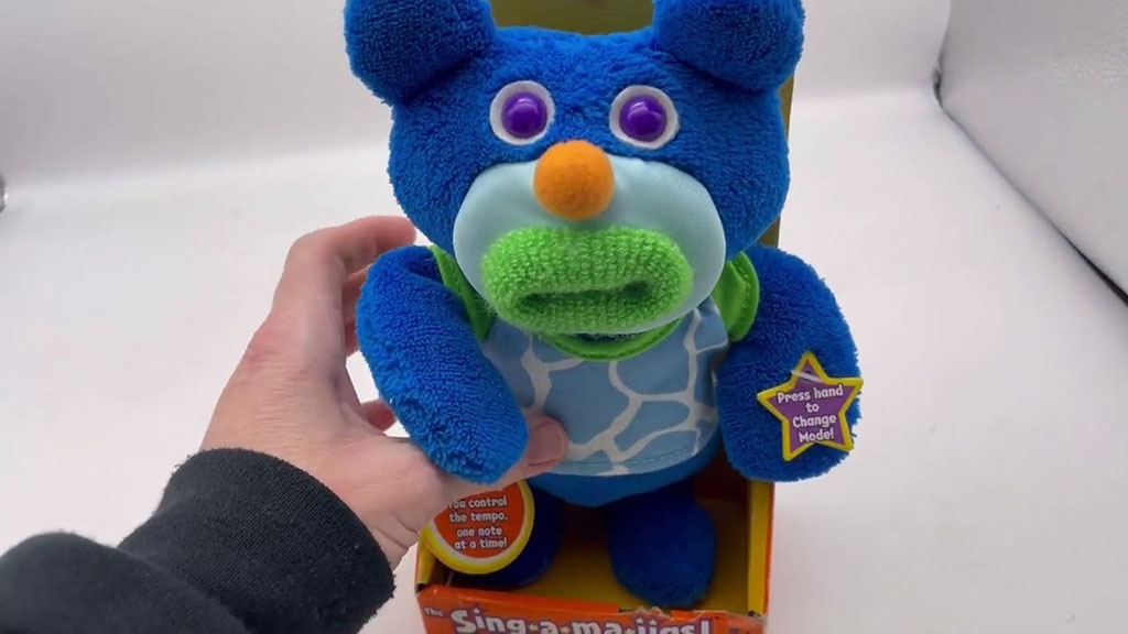 Hand holding a blue interactive plush bear toy with purple button eyes, green muzzle, orange nose, and a patterned vest, displayed in its retail packaging.