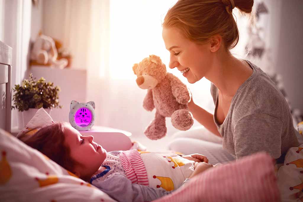 Mother smiling and playing with a teddy bear while her baby lies in bed during a calm bedtime routine