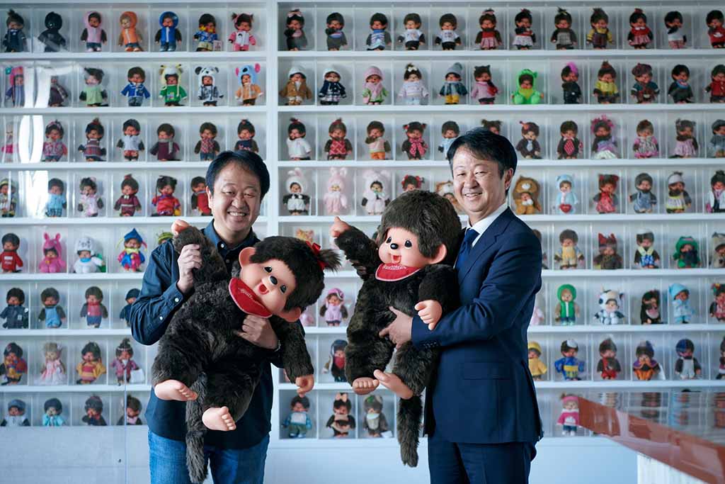 Two adult men standing indoors, smiling and holding large plush monkey dolls, with a full wall of small collectible dolls displayed in clear compartments behind them, creating a colorful toy collection backdrop.