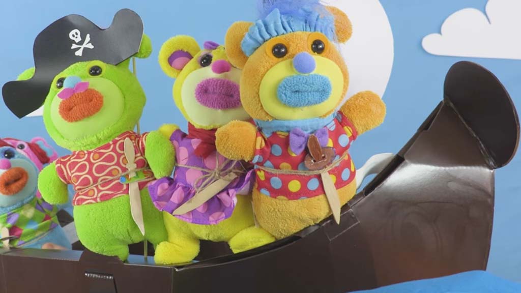 Three colorful plush bear characters dressed in pirate-style costumes, standing together inside a toy pirate ship against a blue sky background.