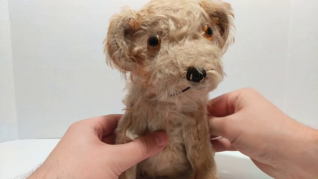 Hands gently holding a small beige plush dog with curly fur, button eyes, and a stitched nose, shown against a plain white background.