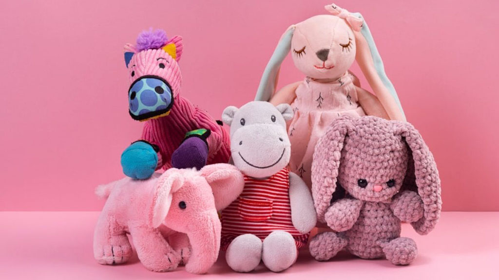 Assorted pastel plush toys arranged on a pink background, including a knitted bunny, soft rabbit doll, hippo in striped outfit, pink elephant, and a colorful zebra plush.