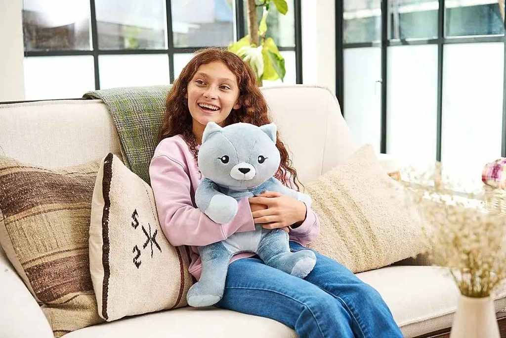 Smiling young girl sitting on a sofa while hugging a blue weighted stuffed animal, creating a sense of comfort, relaxation, and emotional security in a bright living room.
