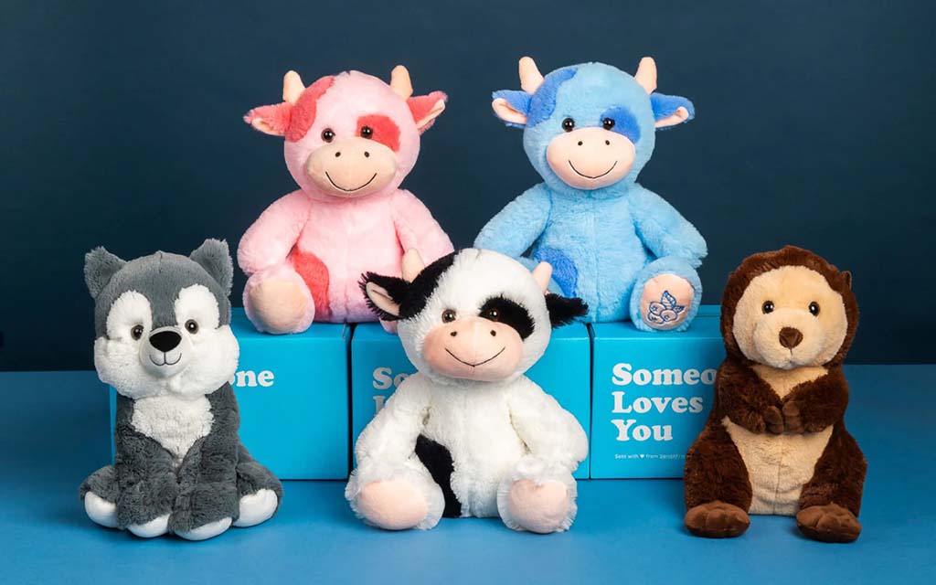Six plush animal toys arranged in two rows against a blue background, including pink and blue cows, a black-and-white cow, a gray wolf, and a brown monkey, displayed with blue gift boxes that read ‘Someone Loves You.