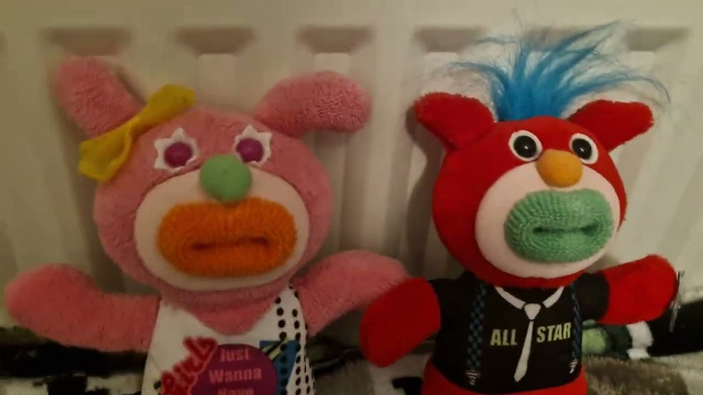 Two colorful monster plush toys sitting side by side on a soft surface—one pink with a yellow bow and polka-dot outfit, the other red with blue spiky hair wearing a black ‘All Star’ shirt—both featuring large stitched eyes and textured noses.