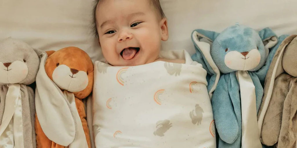 Smiling baby lying on a bed between soft plush comforter toys, showing comfort, security, and gentle sensory companionship.