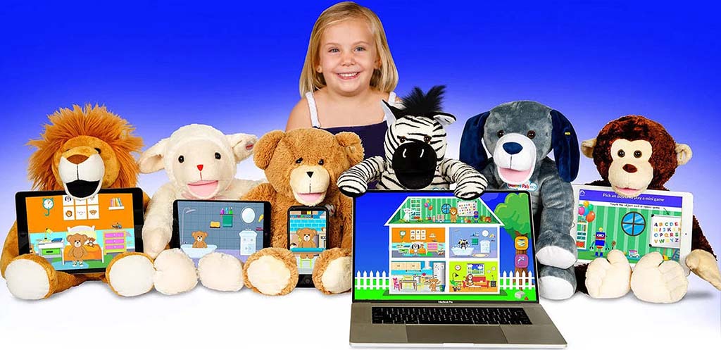 Smiling child seated behind a row of interactive plush animals, including lion, lamb, teddy bear, zebra, dog, and monkey, each paired with tablets or a laptop displaying educational learning apps