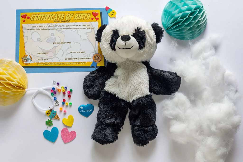 Flat lay of a panda teddy bear making kit featuring an unstuffed black-and-white panda plush, loose polyester stuffing, a colorful certificate of birth, beads, heart-shaped felt pieces, and craft accessories on a white background.