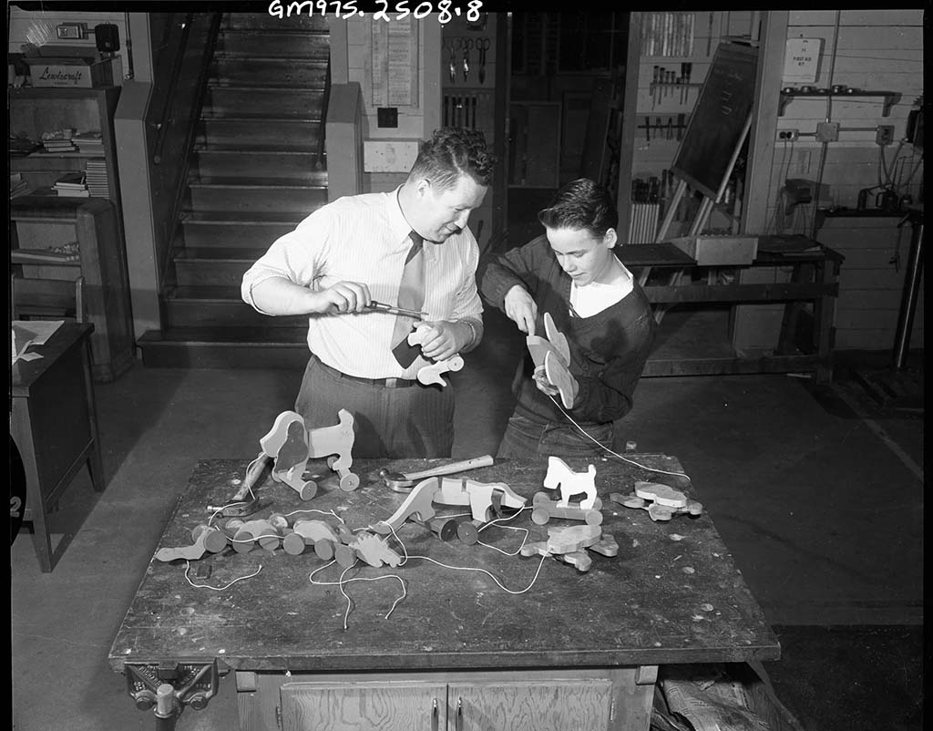 Black-and-white historical photo of an adult and a child working together at a workshop table, assembling wooden pull toys with hand tools, showing an early toy-making process in an educational setting.