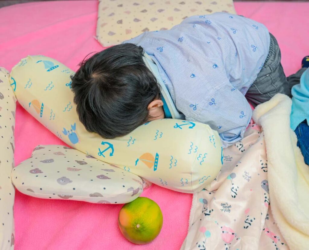 Young child sleeping face down on a pink bed, resting on a soft patterned baby pillow, surrounded by blankets and cushions.