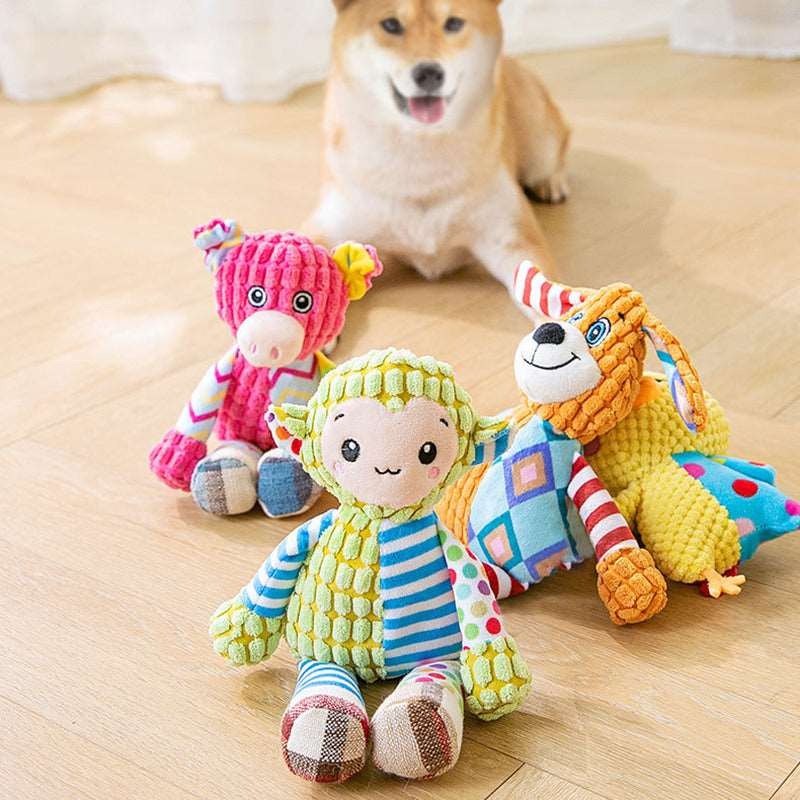 Three colorful plush dog toys with textured fabric and patchwork patterns placed on a wooden floor, with a small dog sitting in the background watching them.