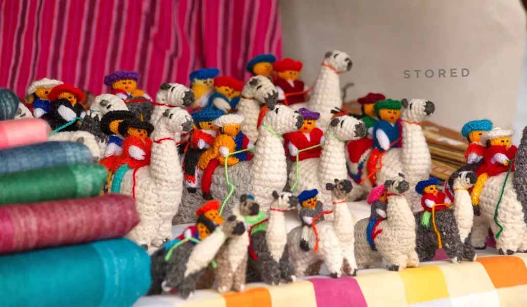 Colorful handmade crocheted stuffed animals arranged in rows on a shelf, featuring small llamas and figures wearing bright hats, with folded fabrics stacked beside them.