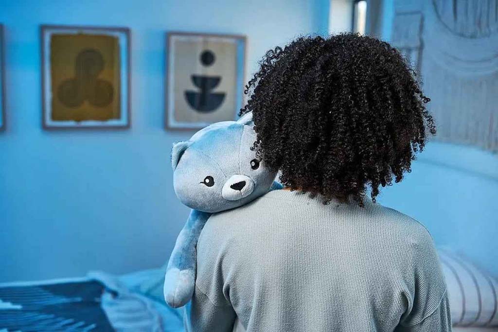 Person holding a blue weighted teddy bear over their shoulder in a softly lit bedroom, conveying comfort and emotional support.