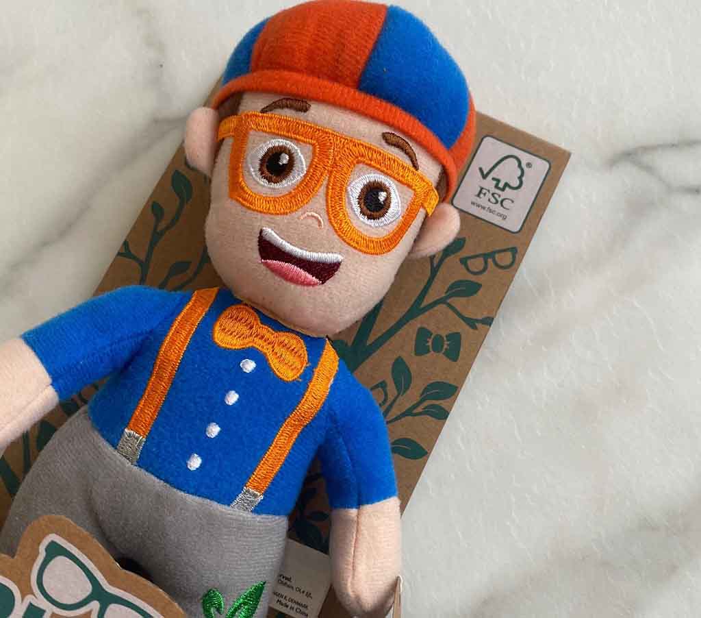 Plush character doll wearing a blue outfit with orange suspenders, orange glasses, and a matching cap, displayed on eco-friendly FSC-certified packaging.
