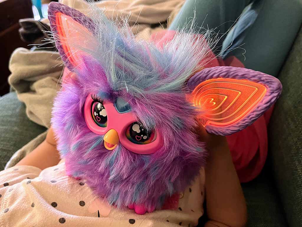 Colorful interactive plush toy with fluffy purple and blue fur, glowing LED ears, large expressive eyes, and a small beak, resting on a child’s lap in a cozy indoor setting.