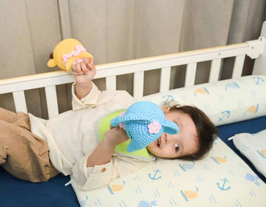 Baby lying in a crib, holding two soft crocheted plush toys while playing on a patterned mattress.