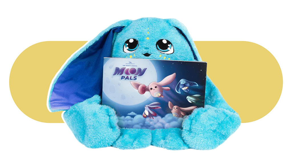 Blue plush bunny holding a ‘Moon Pals’ storybook, featuring large embroidered eyes and floppy ears, shown against a soft yellow background.