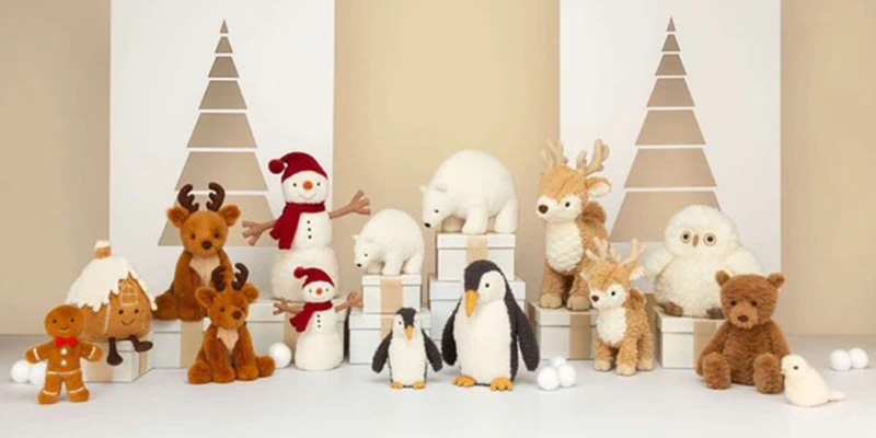 Festive holiday display of plush toys including reindeer, snowmen, penguins, polar bears, owls, and a teddy bear arranged in a winter Christmas scene with minimalist tree backdrops.