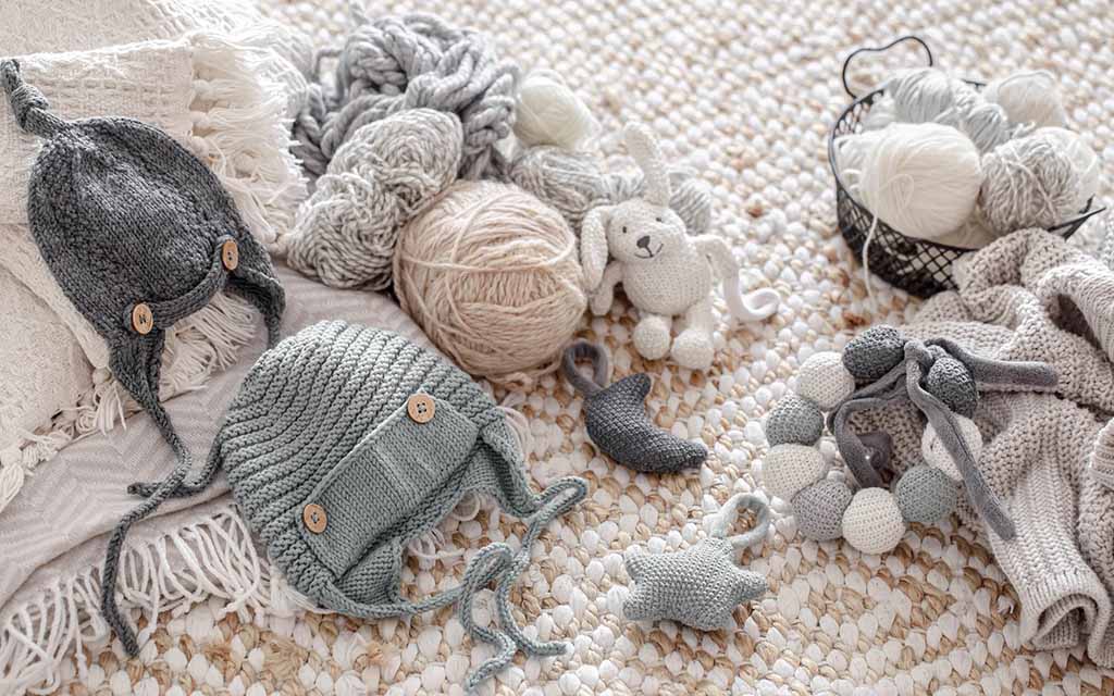 Handmade knitted baby toys and accessories including soft hats, crochet animals, yarn balls, and textured rattles arranged on a neutral woven rug.