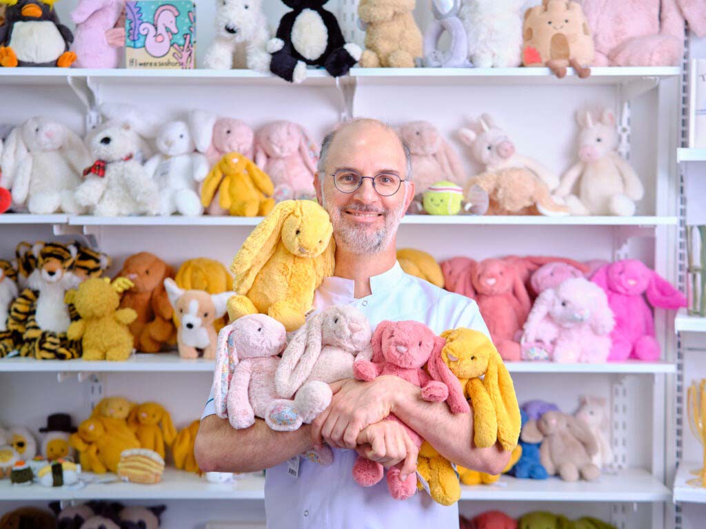 A smiling man standing in front of shelves filled with colorful plush toys, holding several soft bunny plushies in pastel yellow, pink, and beige tones.