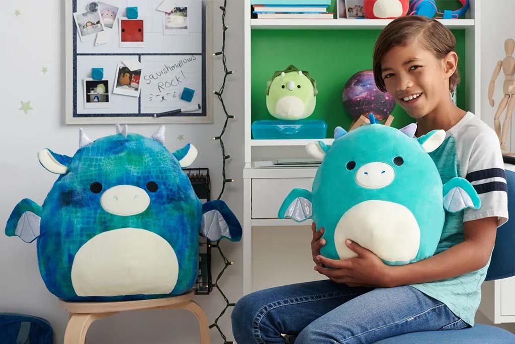 A smiling child sitting indoors while holding a large soft plush toy, with another oversized plush displayed beside them in a cozy bedroom setting.