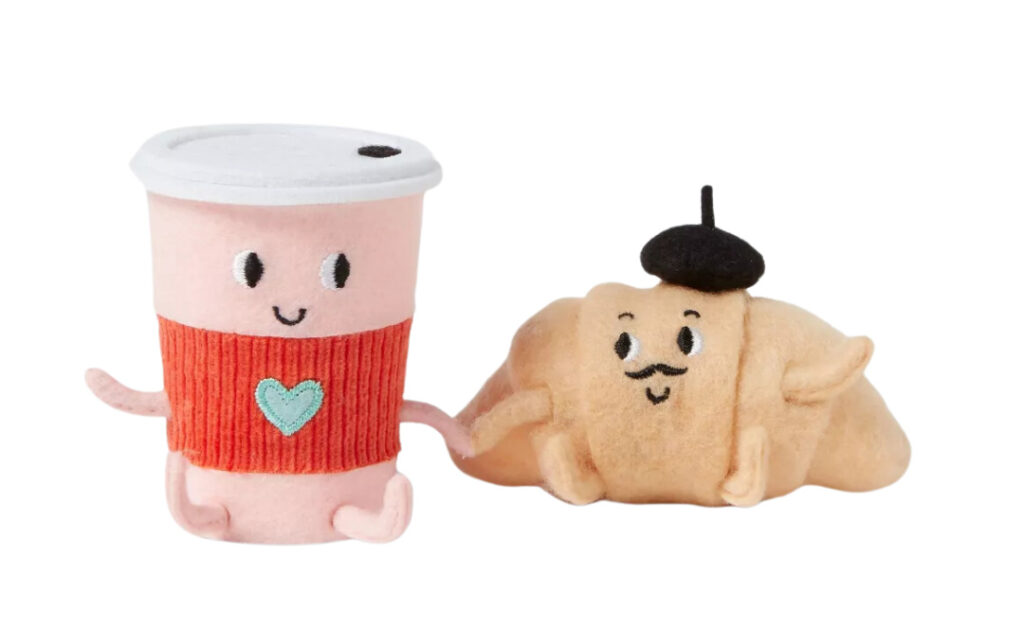 A plush coffee cup and a plush croissant holding hands, both with smiling embroidered faces; the coffee cup wears a red sleeve with a small heart, and the croissant has a tiny black beret.