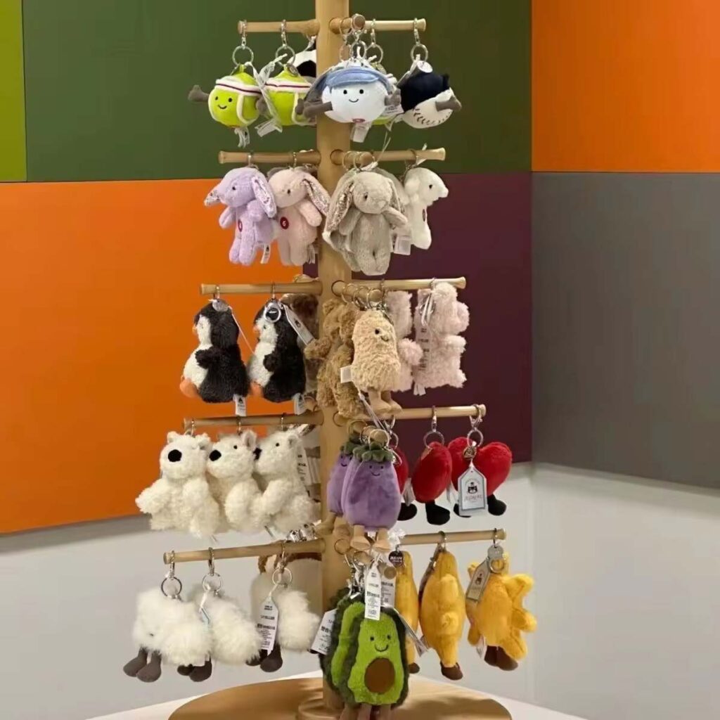 A rotating wooden display stand filled with assorted Jellycat-style plush keychains, including bunnies, penguins, sheep, avocado characters, bananas, suns, hearts, and other mini plush designs, arranged in rows against a colorful wall background.