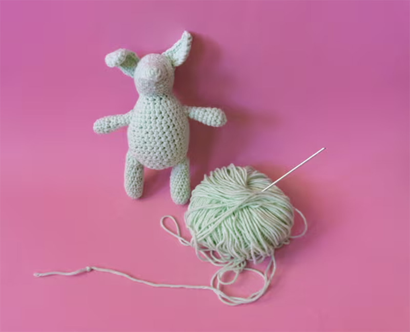 Handmade crocheted plush bunny displayed on a pink background beside a ball of yarn and needle, illustrating amigurumi toy craftsmanship, soft yarn materials, and the creative process behind handmade and custom plush toy production, ideal for showcasing textile techniques and artisanal plush design inspiration.