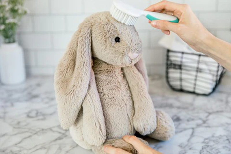 A beige bunny plush toy being gently brushed with a handheld brush on a marble countertop, showing cleaning and care of a stuffed animal.