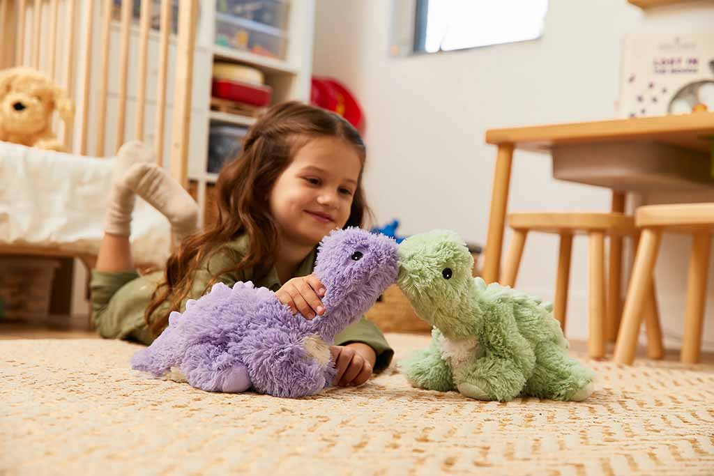 A young girl lies on a carpeted floor in a cozy bedroom, happily playing with two soft plush dinosaur toys—one purple and one green—creating a warm and imaginative playtime scene.