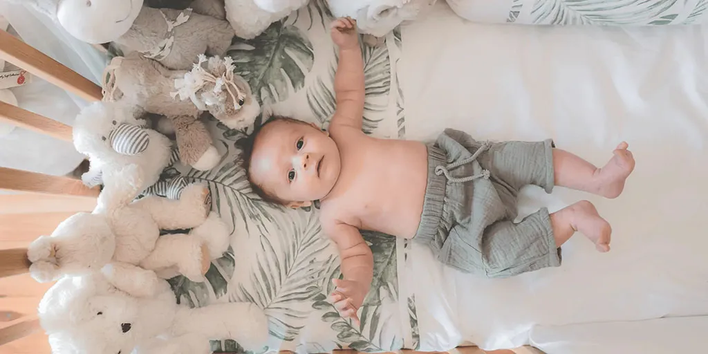 A baby lies on a leaf-patterned crib sheet surrounded by soft plush animals, looking up while resting comfortably in the crib.