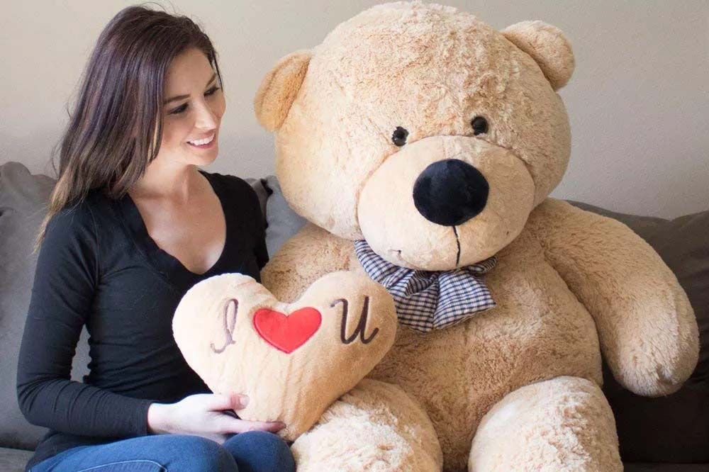 A woman sitting beside an oversized teddy bear holding a heart-shaped pillow, illustrating large plush toy scale, soft fabric texture, and emotional gifting appeal for premium stuffed animals in retail and wholesale markets.