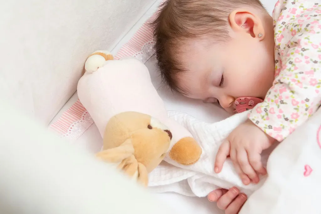 A baby sleeps peacefully in a crib, holding a blanket beside a soft plush toy for comfort.