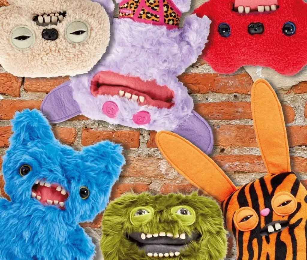 A collection of colorful Fuggler plush monsters with quirky faces, mismatched eyes, goofy teeth, and textured fur arranged against a rustic brick wall background.
