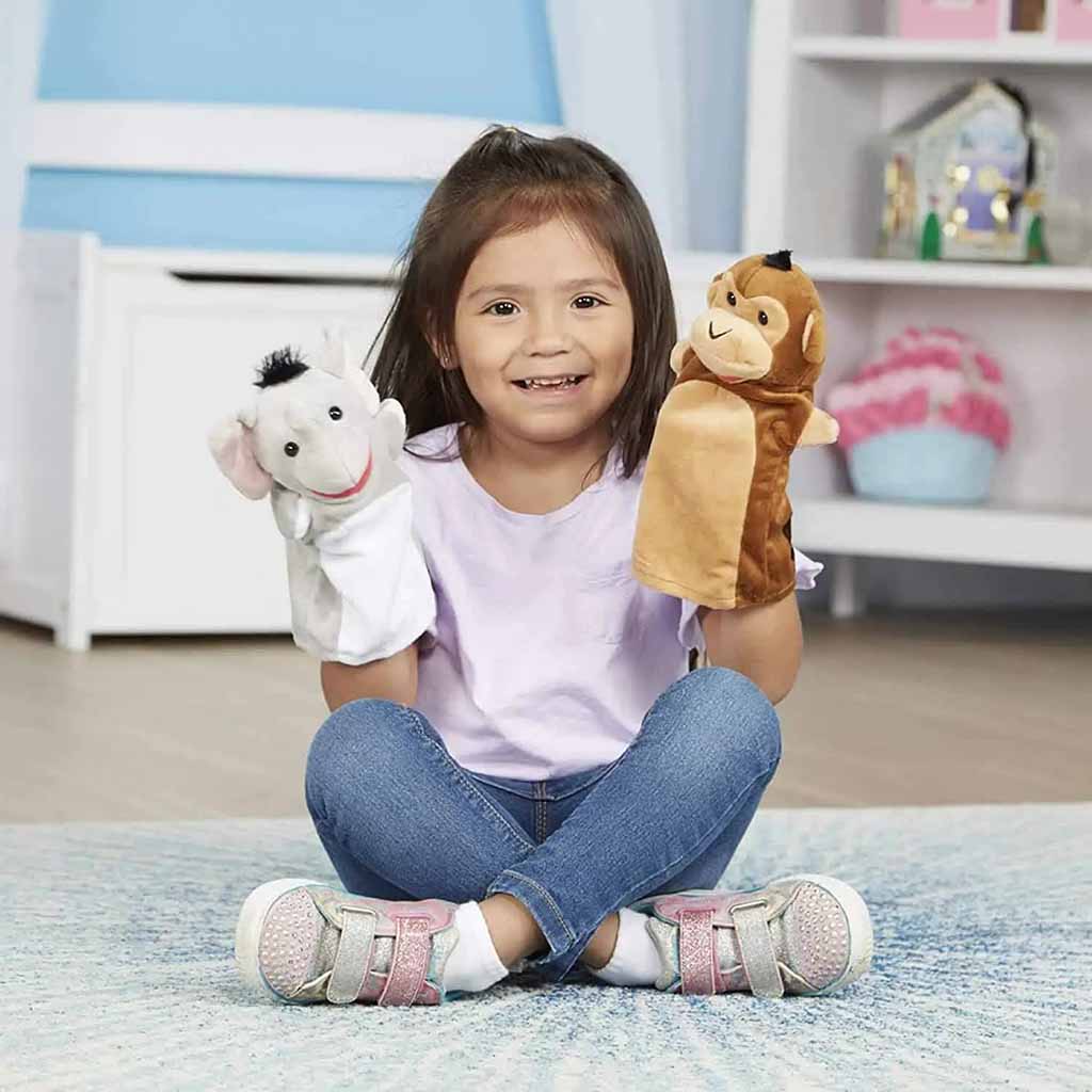 A smiling young girl sitting cross-legged on the floor while holding two plush hand puppets, one white mouse and one brown monkey, in a bright playroom setting.