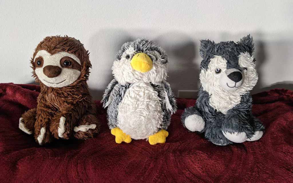 Three plush toys—a brown sloth, a gray-and-white penguin with a yellow beak, and a gray wolf—sitting side by side on a red blanket against a light-colored wall.
